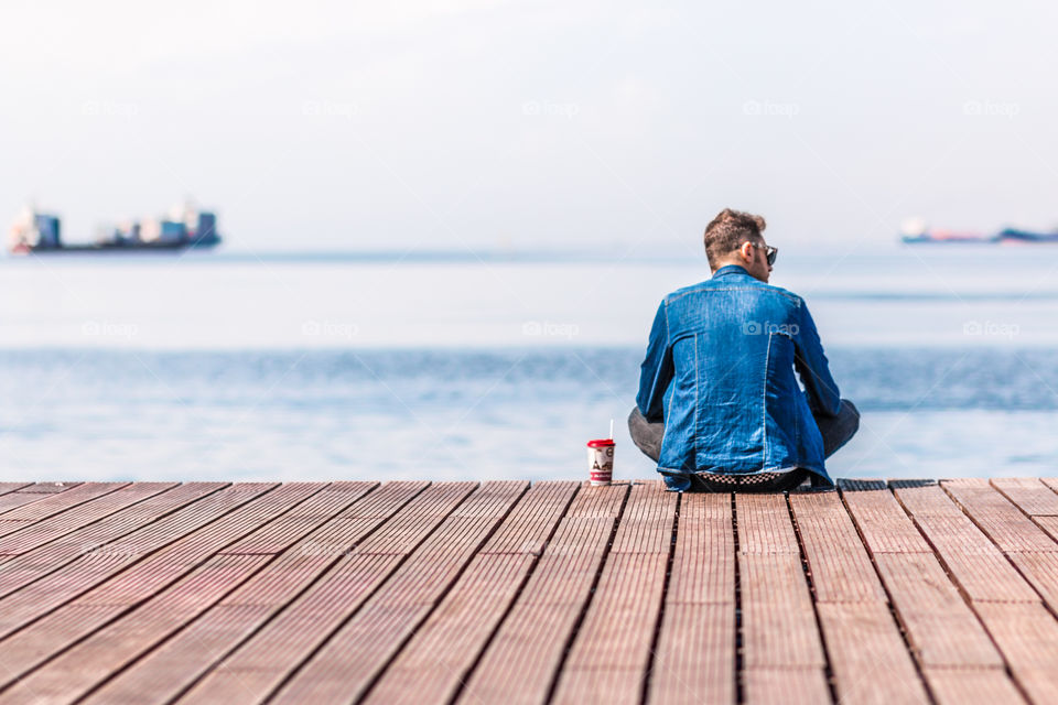 Man Wearing Denim Jeans Clothes Sitting On A Dock And Looking At Blue Sea
