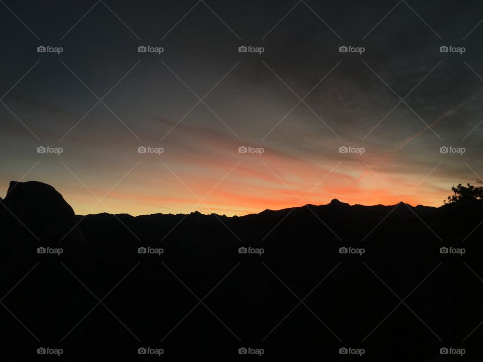 A silhouette view of the Yosemite Mountains at early dusk where the mountains themselves are almost completely darkened and the sky has just begun to illuminate vibrant pink and orange tones in the clouds above. 