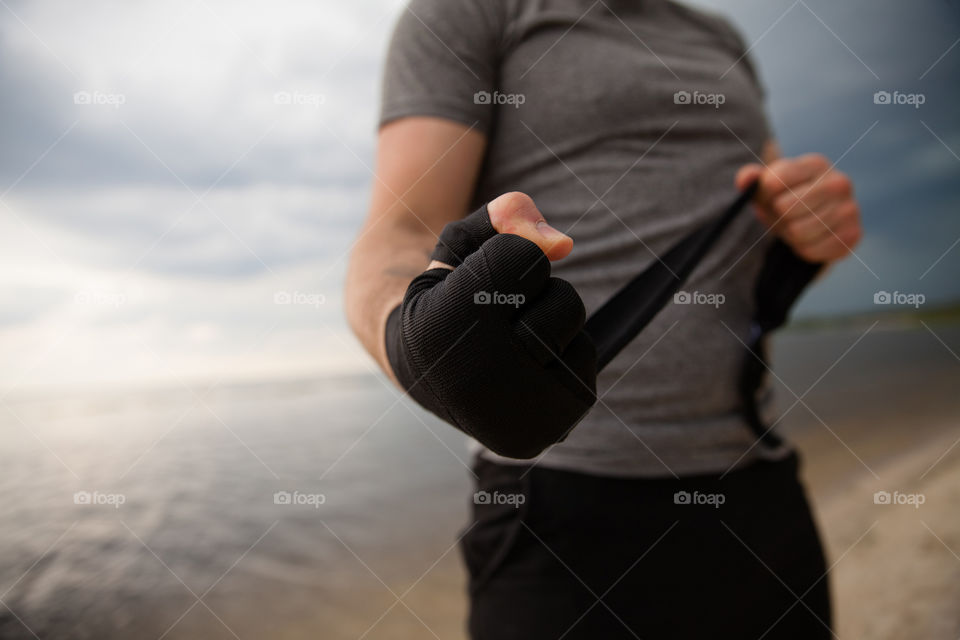 an athlete conducts a warm-up on the seashore.  a guy in a gray T-shirt, black shorts, rewinds his hands with a bandage under boxing gloves