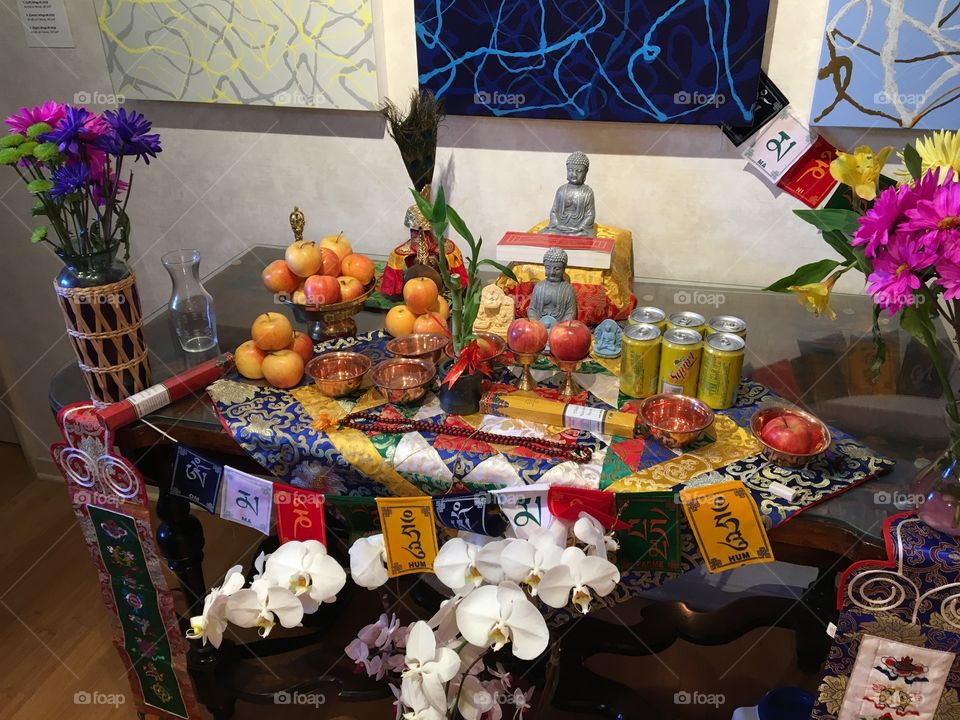 Fruit, flowers, incense, Buddha statues, and prayer beads stand ready on an altar during a sand mandala ceremony.