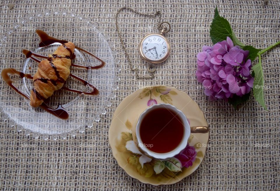 Fresh croissant with brewed tea in antique teacup accented with vintage pocket watch and hydrangea 