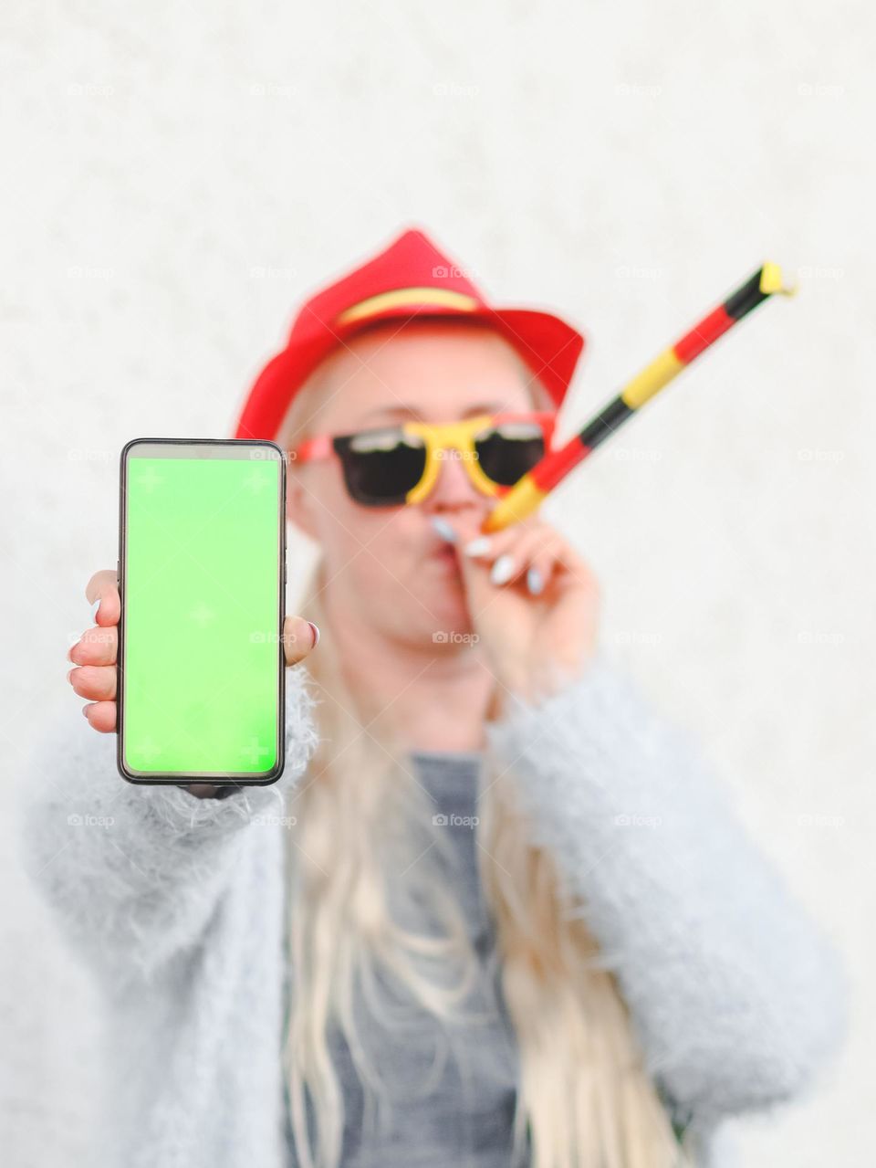 Portrait of a beautiful young caucasian blonde woman in a Belgian flag hat, sunglasses blowing a whistle and holding a mobile phone in her outstretched left hand with a green empty screen on a white wall, side view close-up.