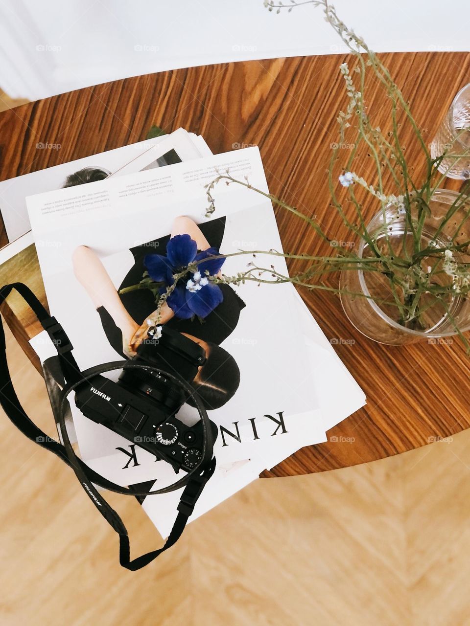 A stack of magazines and a camera lay on a wooden table in a bright room, nobody 