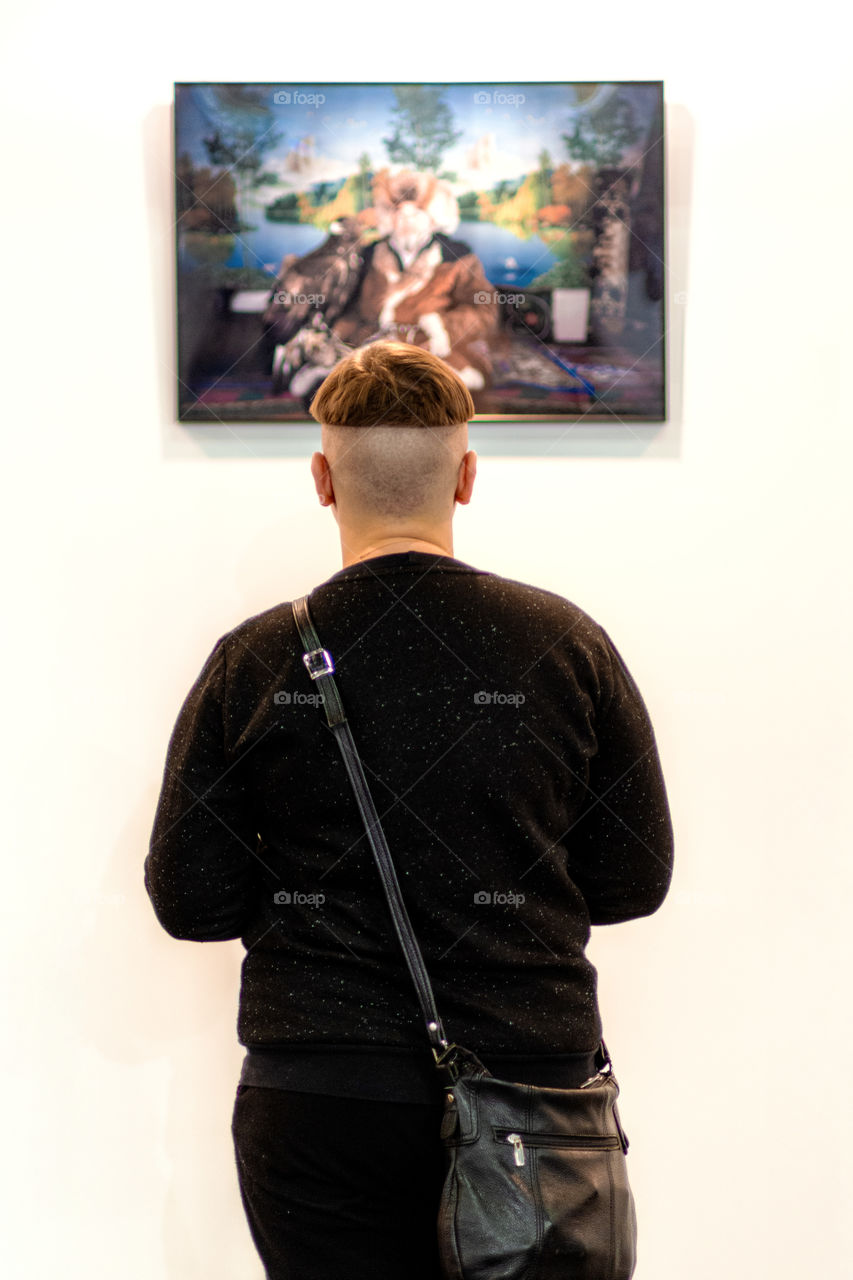 A man with an unusal haircut stands on a white wall and looks a photograph. Rear view.