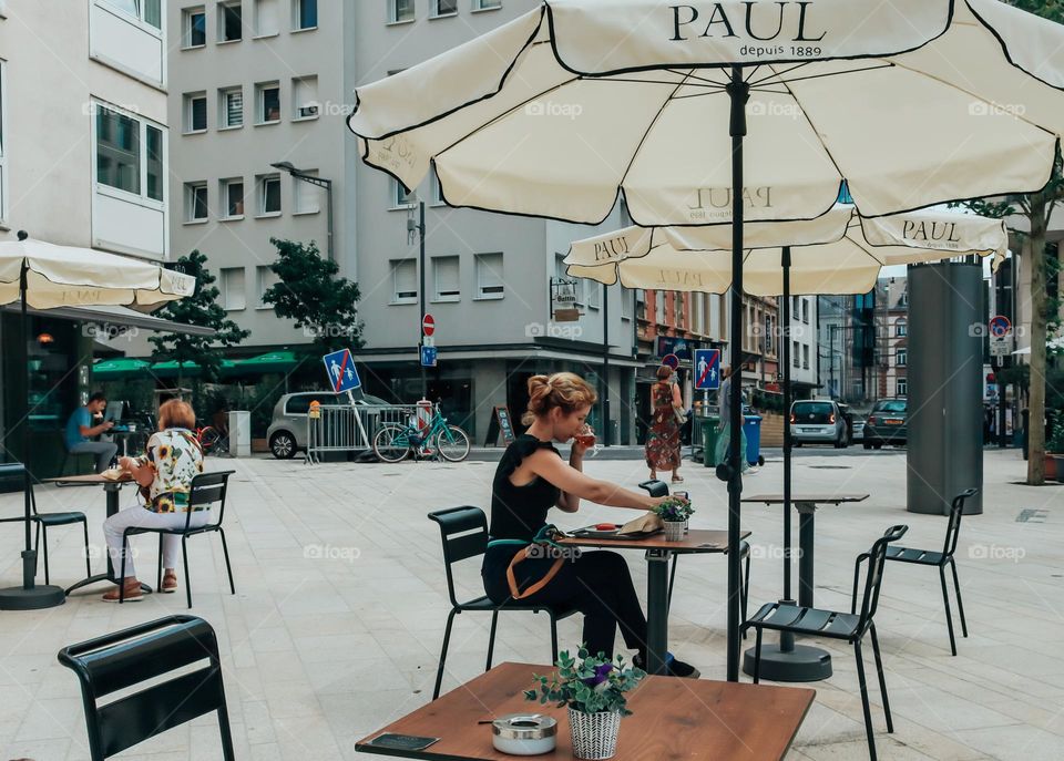 One young recognizable caucasian girl sits alone at a street cafe table and drinks coffee on a summer morning in the center of Luxembourg city, close-up side view.