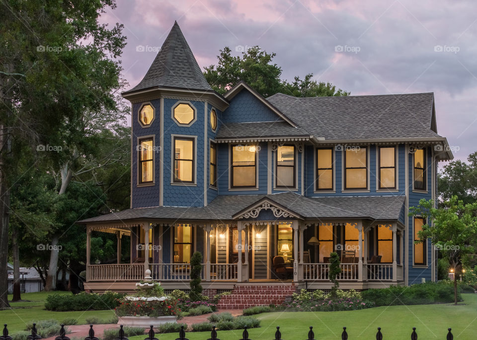 historic Victorian style house with warm golden interior lights, green grass, and red brick walkway in front of a purple and pink evening sky