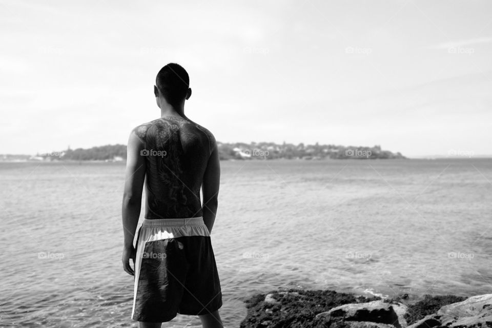 Man with a full back tattoo facing the ocean