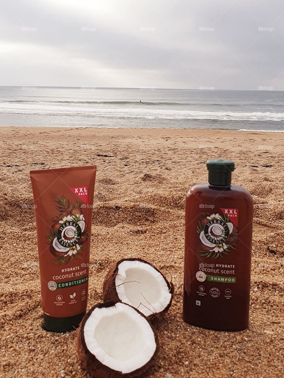 Product composition for Herbal Essences Coconut scent shampoo and conditioner placed on sand with a coconut split in two and the sea as background
