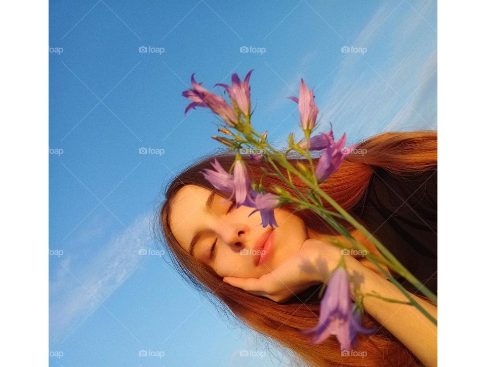 It is a photo of a young woman in the field. She has red (ginger) hair. She has brown eyes. Her face is partly coverd with Campanula patula or spreading bellflower