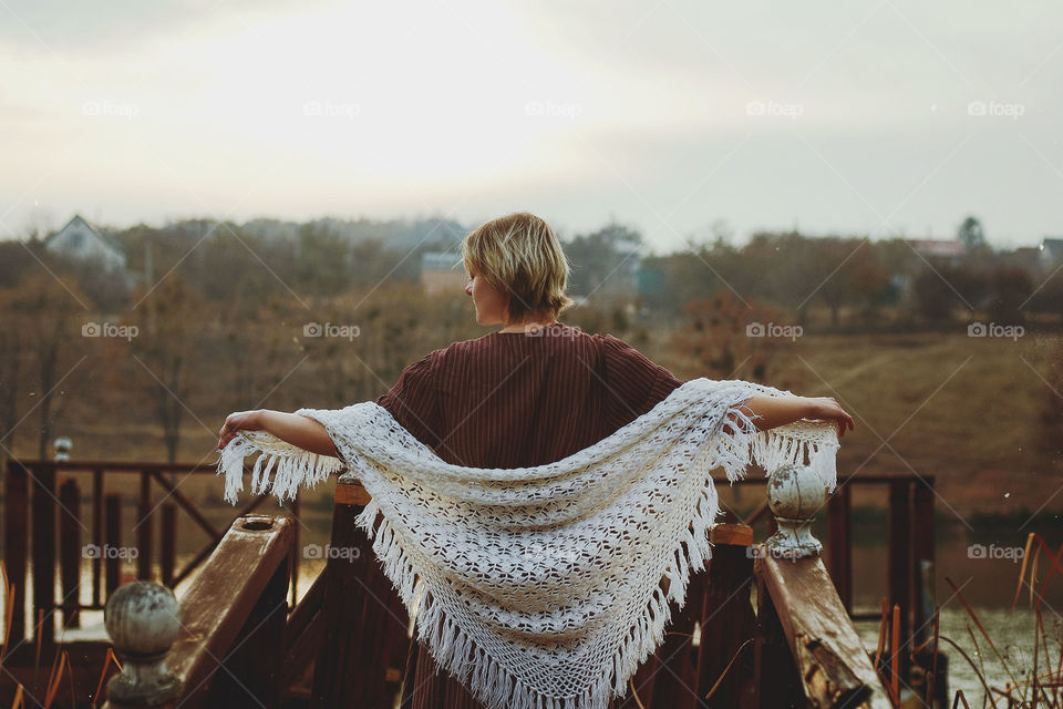 Beautiful portrait of young sad woman standing on the bridge near river with big white scarf. Autumn cold season.