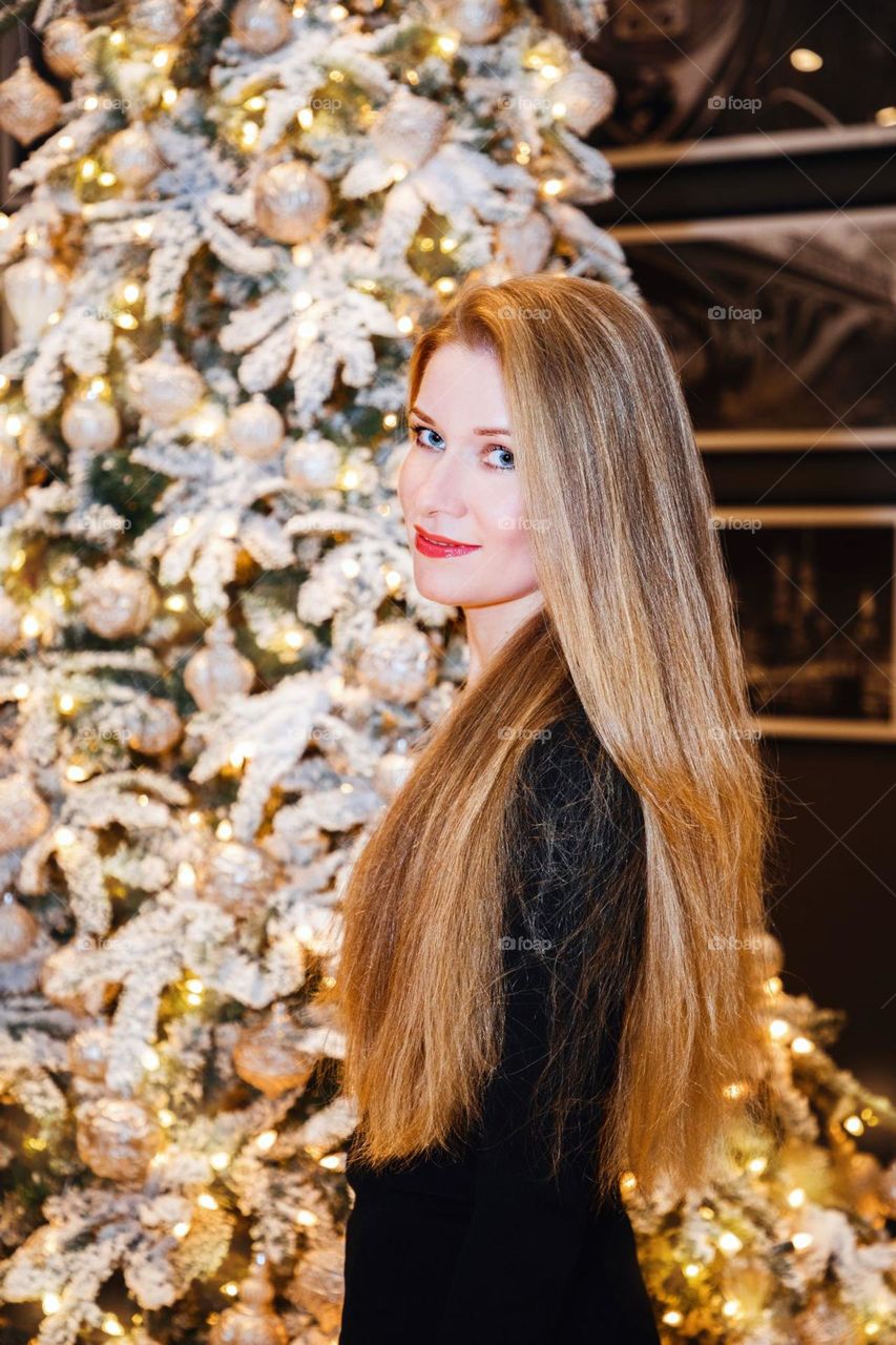 Blonde female with beautiful long hair near richly decorated tree; she is looking at us