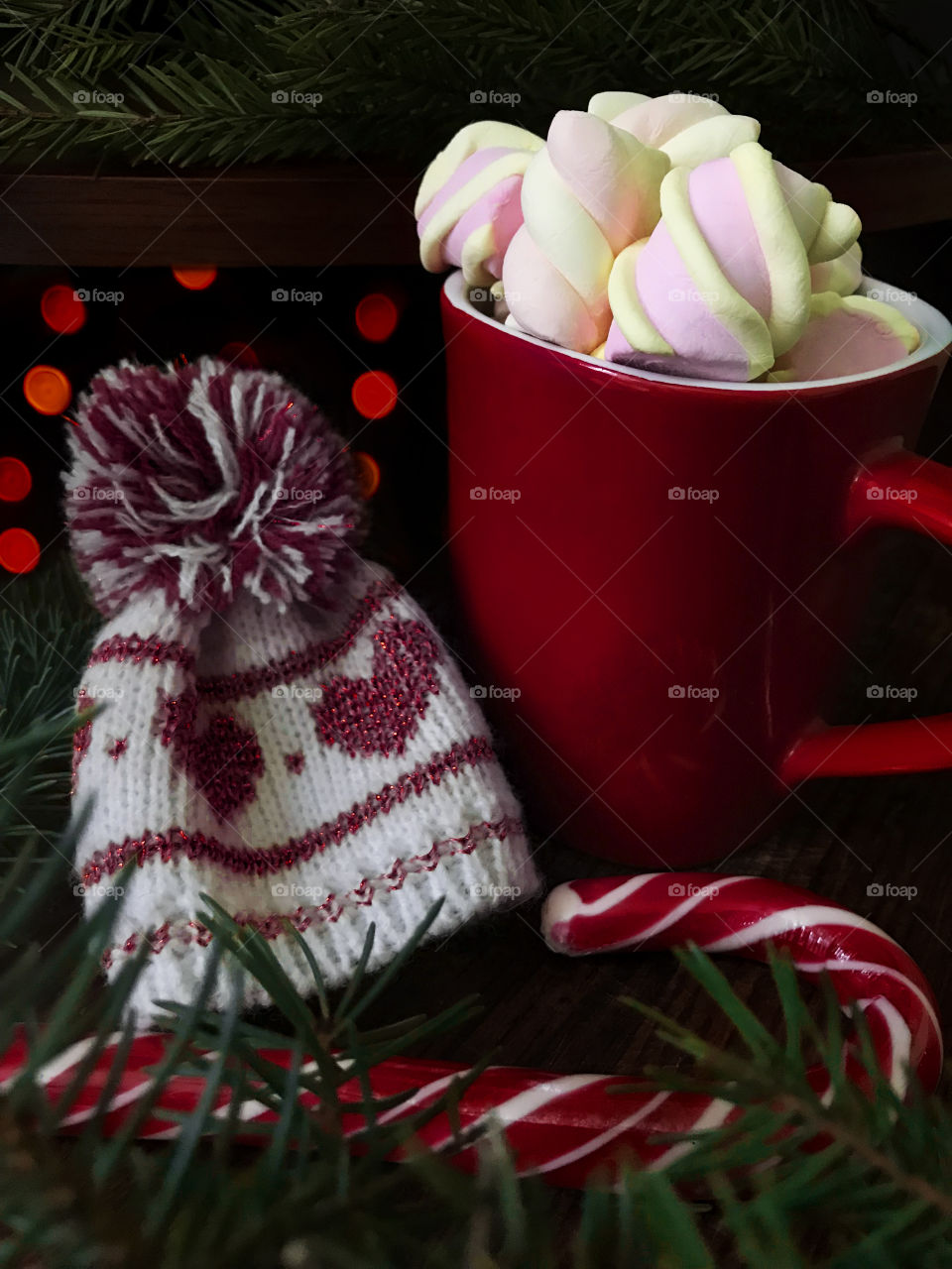 Hot coffee/cocoa with marshmallows in red cup nearby candy cane, fir tree branch and winter hat