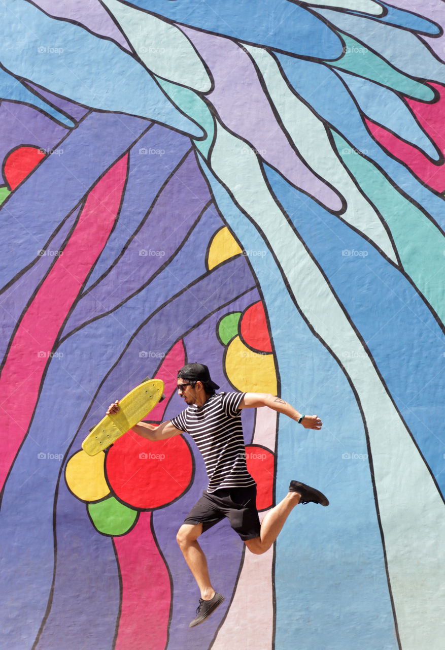 Side view of a young man jumping high with his cruiser skateboard in his hand in front of a colorful graffiti.