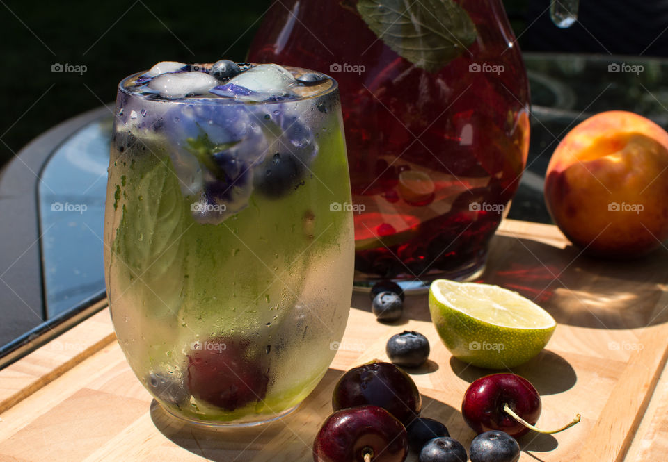 Beautiful glass of cherry, lime and mint flavored water with floral ice cubes on wood table in garden with colorful pitcher of blueberry peach homemade drink garnished with fresh berries, aromatic herb and fruit ingredients 