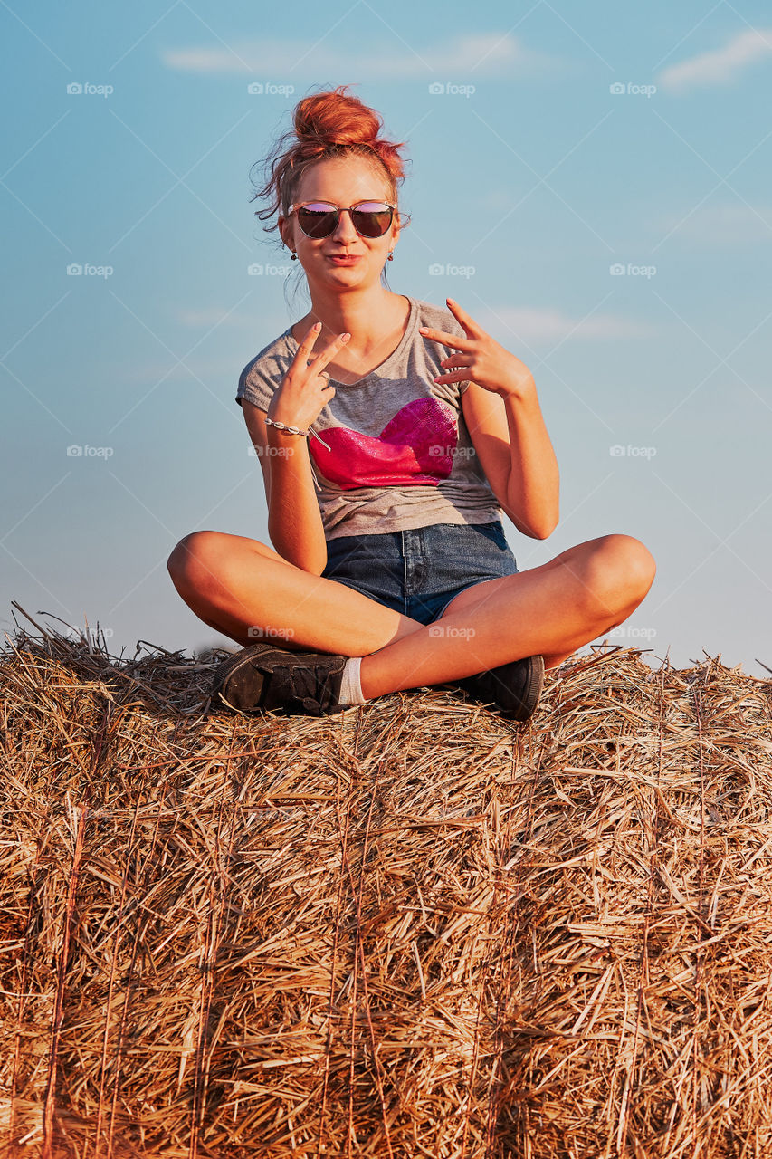 Happy smiling teenage girl sitting on a hay bale at sunset making V sign gesture enjoying summer vacations in the countryside. Candid people, real moments, authentic situations