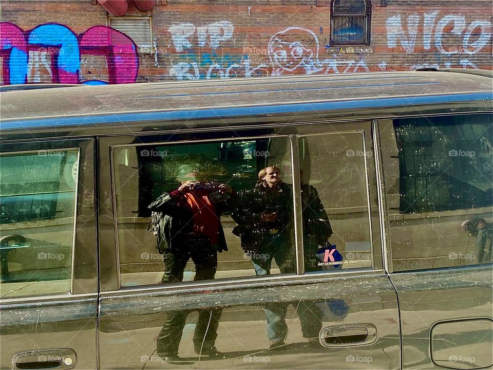 Timothy & Petra reflected in a mirror tinted large car window on “Waverly St” in “Fort Greene”, Bklyn as they are walking across the street. 2022. Hypnotic Productions