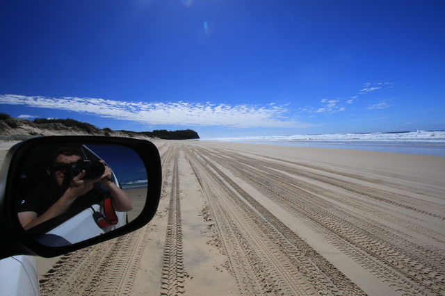 Fraser Island beach photo including photographer reflection in the car wing mirror. Car tracks in the sand.