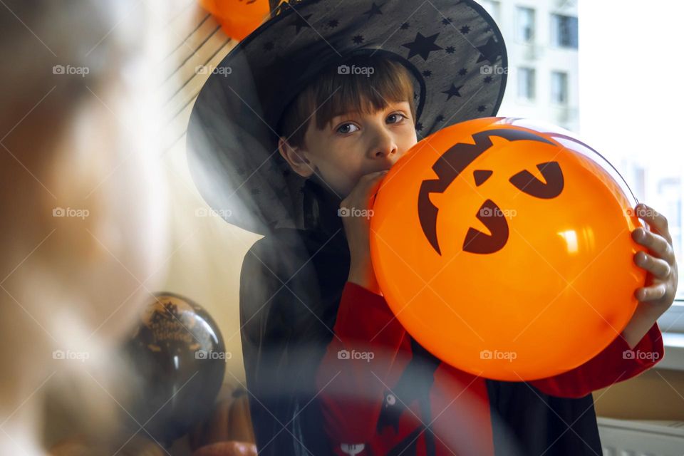 Boy in a black cape and a witch's hat blows a balloon with the image of a pumpkin jack.