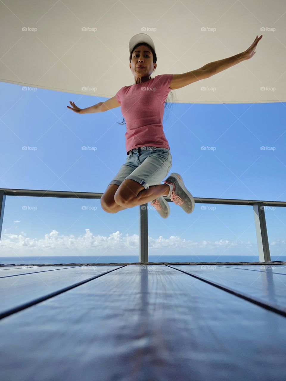 Young adult jumping against blue sky and clouds wearing dark pink shirt with disgusted expression. 