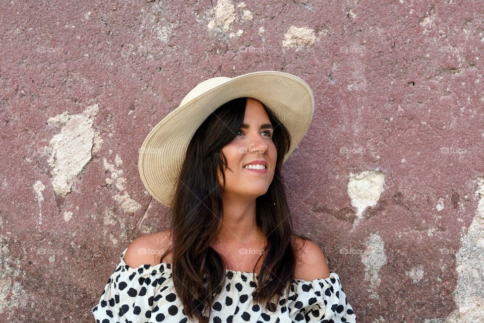 Portrait of beautiful young woman with long dark hair, wearing a hat, standing in front of weathered wall