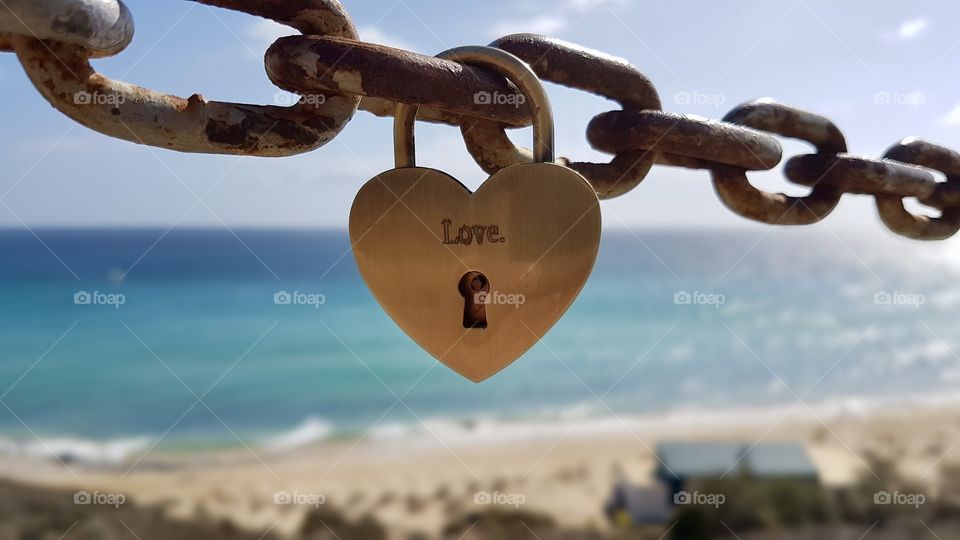 Love is forever, golden heart shaped padlock with the word love hanging on a rusty metal chain, blue turquoise sea in the background  - Guldfärgat hänglås format som ett hjärta med text Love på, hänger på en gammal rostig kedja, hav strand 