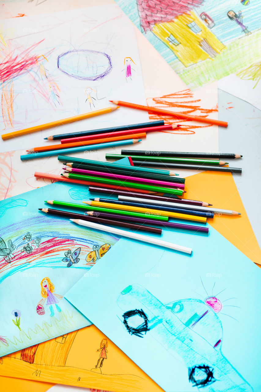 Pencil crayons scattered on desktop filled with colorful childlike drawings of playing children, house, car. Photo from above