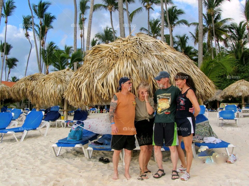 Group of people on beach getting ready for a photo. They're all standing up and nobody is looking at camera. It's really a funny picture. Thatched umbrella & palm trees are in background.