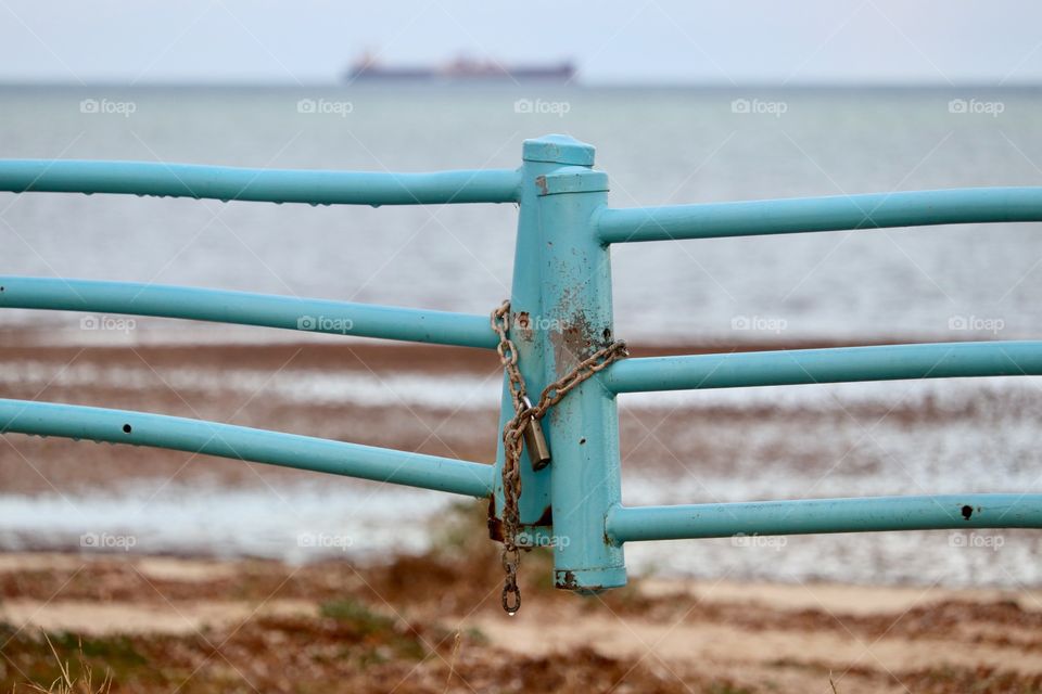 Locked gate to ocean. A turquoise coloured iron metal painted gate with rusty chain and padlock foreground and cargo ship off on the horizon on cloudy dreary day at ocean 