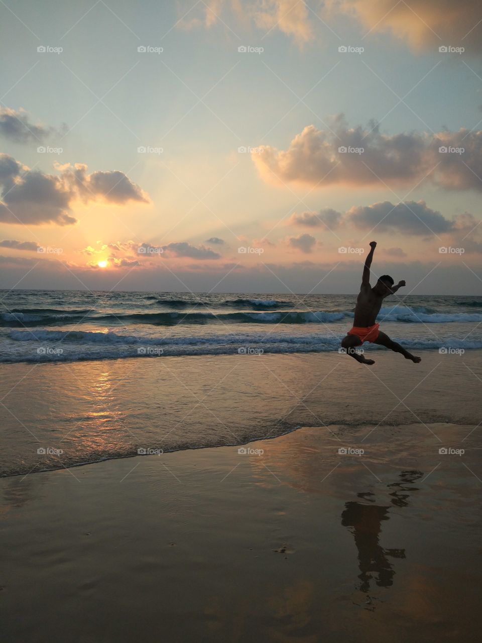 Young man in orange shorts jumping on the seashore at sunset