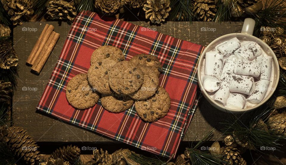 Christmas cookies laid out on a red tartan napkin, with hot chocolate and marshmallows surrounded by a pine cone and green tinsel garland