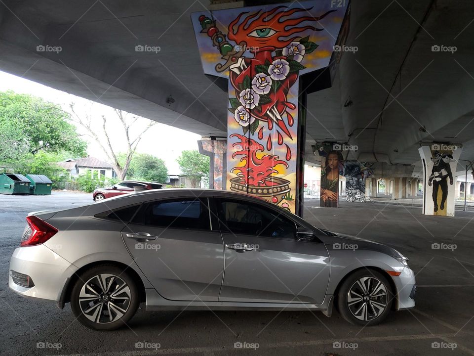 Gray Honda sedan parked in parking lot underneath highway with street art on the highway cement pillars.