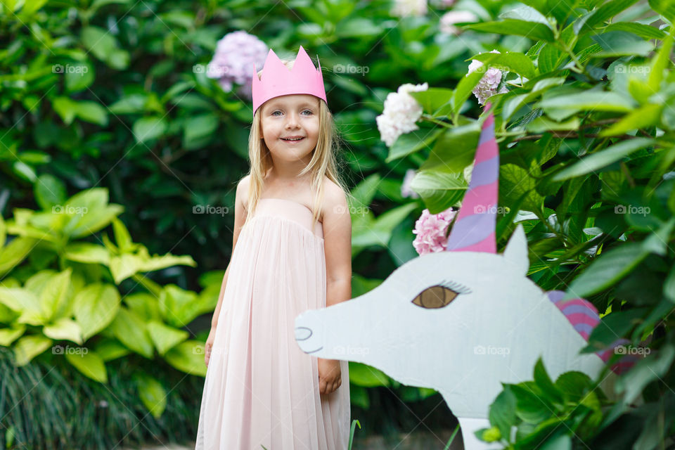 Happy little Caucasian girl with blonde hair and crown standing near unicorn in blooming garden at summer day