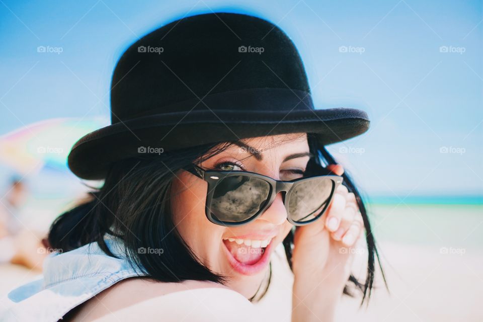 Portrait of attractive brunette woman in bowl hat and retro sunglasses sitting on the bench at summer and smiling with open mouth 