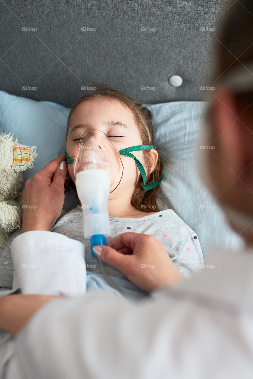 Doctor visiting little patient at home. Child having medical inhalation treatment with nebuliser. Girl with breathing mask on her face. Woman wearing uniform and face mask