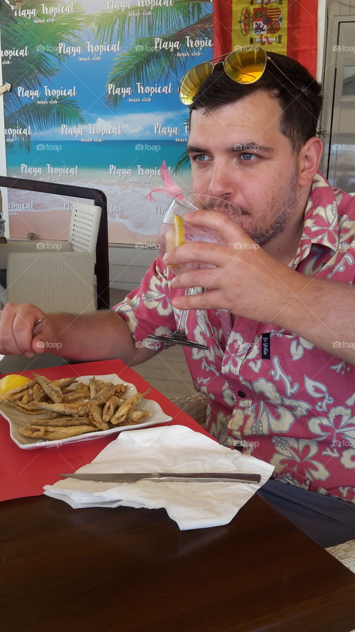 Man Eating Whitebait at a seafood restaurant by the sea and drinking cocktails - bright loud flowery shirt - relaxing on holiday - fish