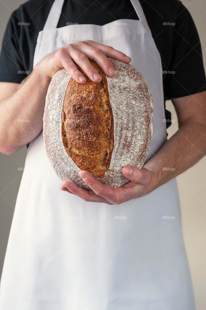 Close-up at bakers hands holding a loaf of sourdough bread in front of him.