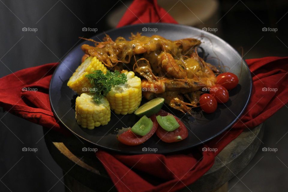 Asian food balado fried prawns with corn chunks, lemon slices and tomatoes served on black plate with red cloth bottom and wooden coaster. Asian style street seafood.