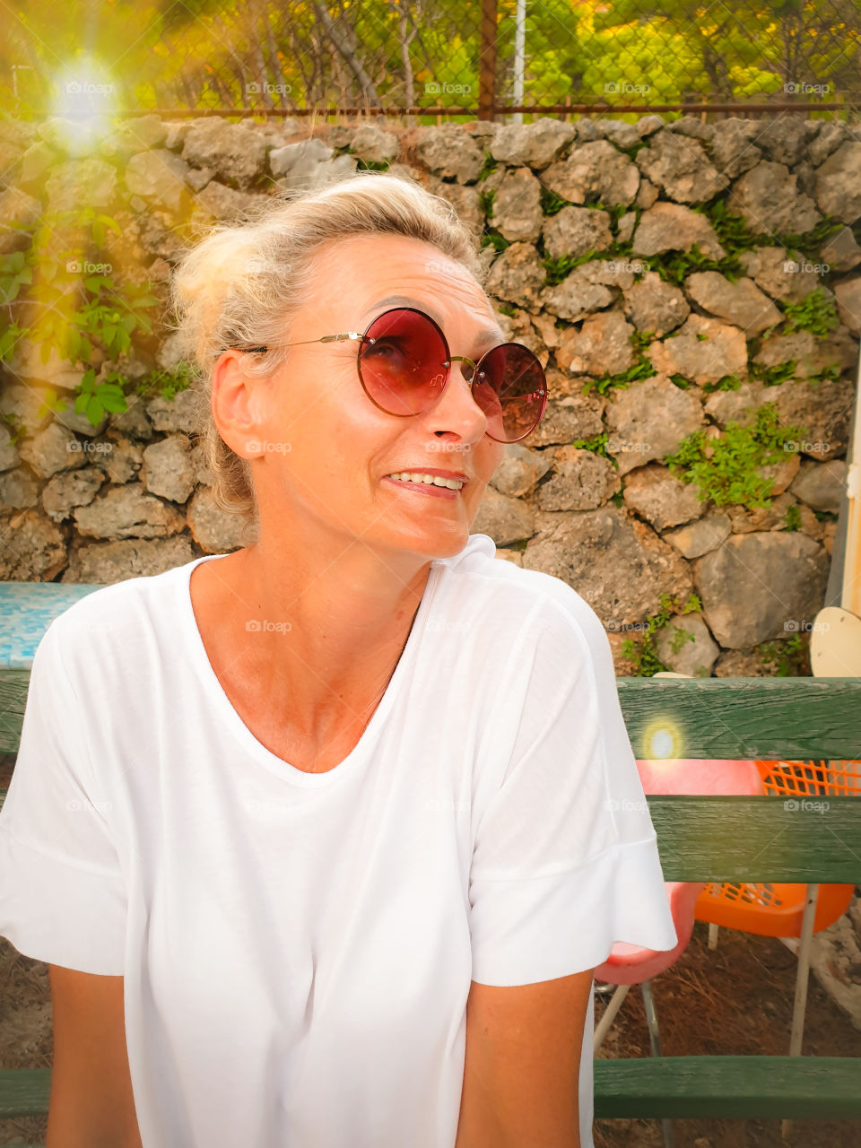 Portrait of a smiling woman in round sunglasses and a white T-shirt against a stone wall.  Lovely aged woman