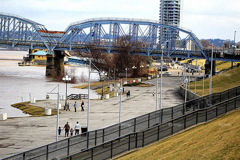 This is a picture of the Ohio River with people walking along and a bridge in the background.