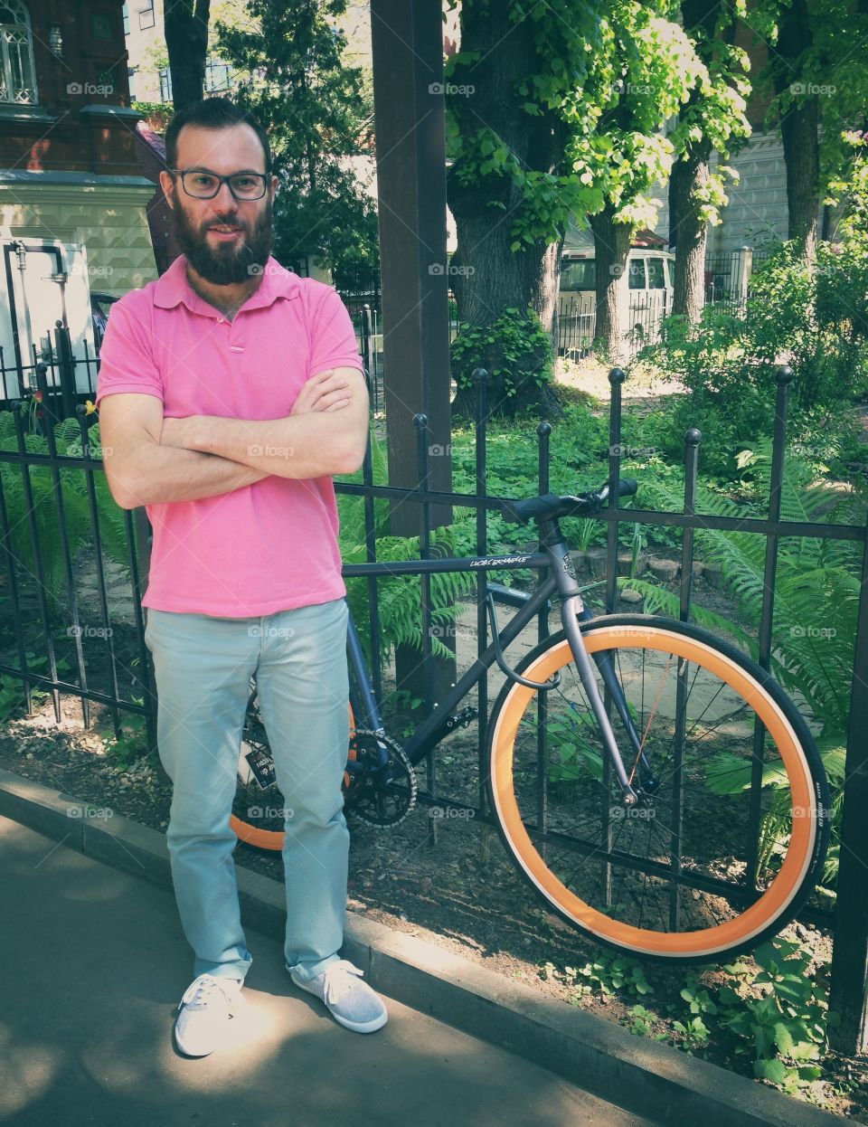Man in pink polo t-short and neon trousers with glasses and beard standing near grey brakeless fixie bike with orange wheels