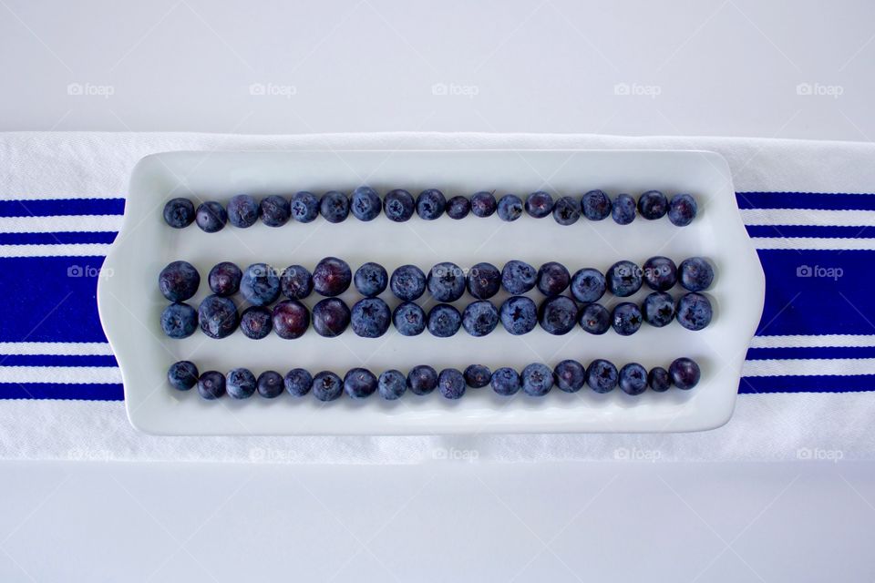 Horizontal flat lay of blueberries on a rectangular white plate, arranged in lines to emphasize the blue stripes on the white dish towel underneath, all on a white background