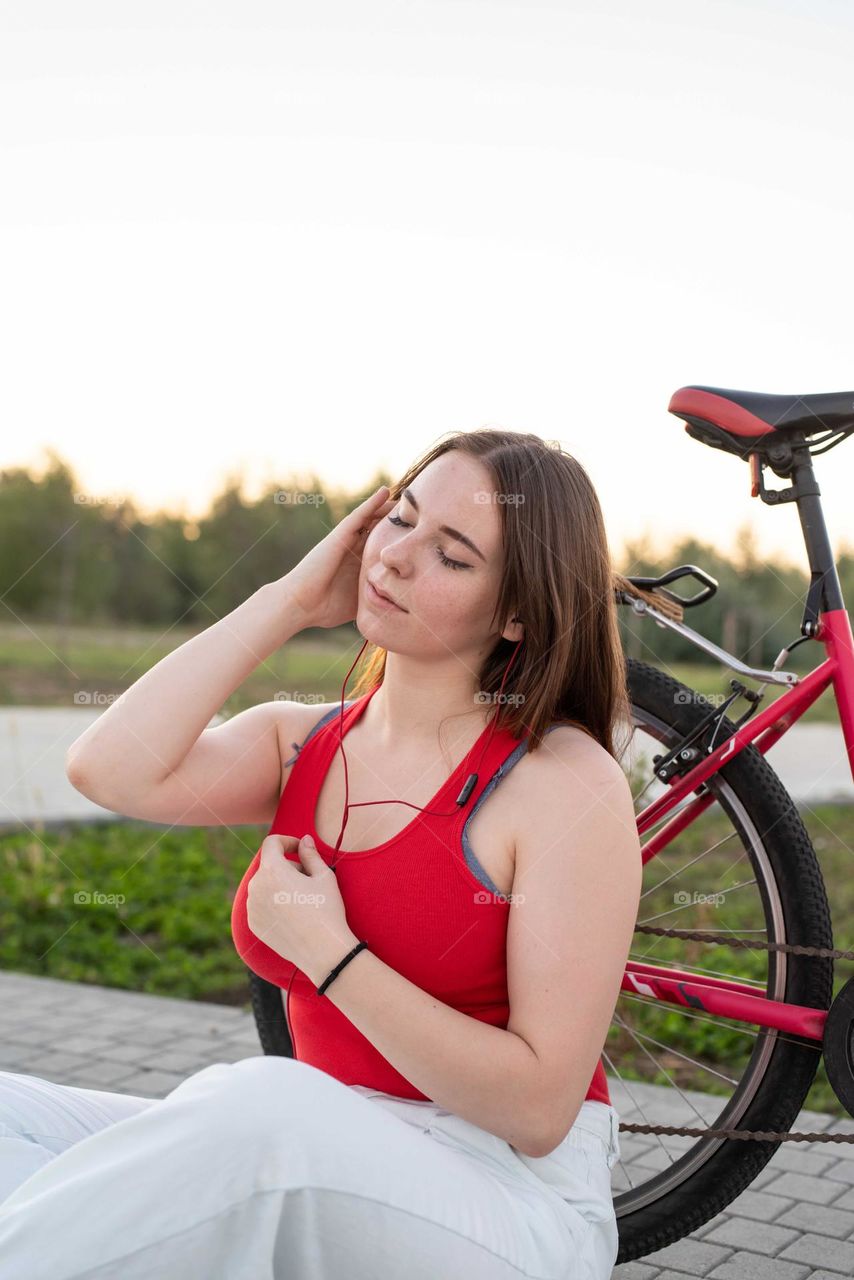 young smiling woman in red shirt and white pants listening to the music standing near her bicycle