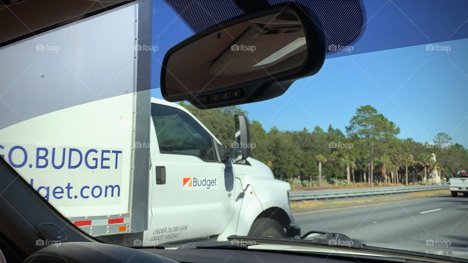 Commuting: inside a vehicle riding on the passenger side seeing a white moving truck passing the vehicle on the highway by green trees.