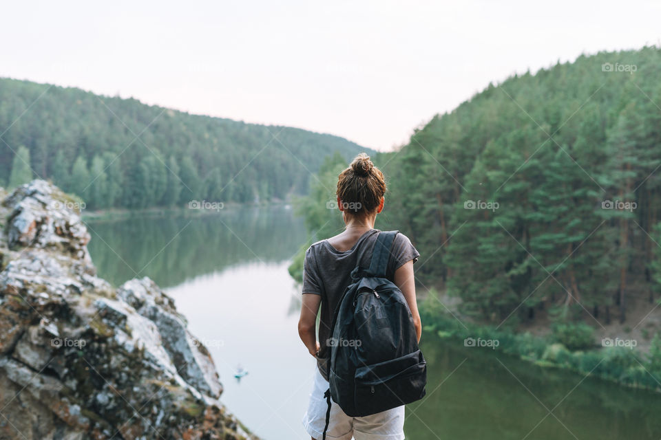 Young slim woman in casual clothes with backpack looks at the beautiful view of mountains and calm river, local travel