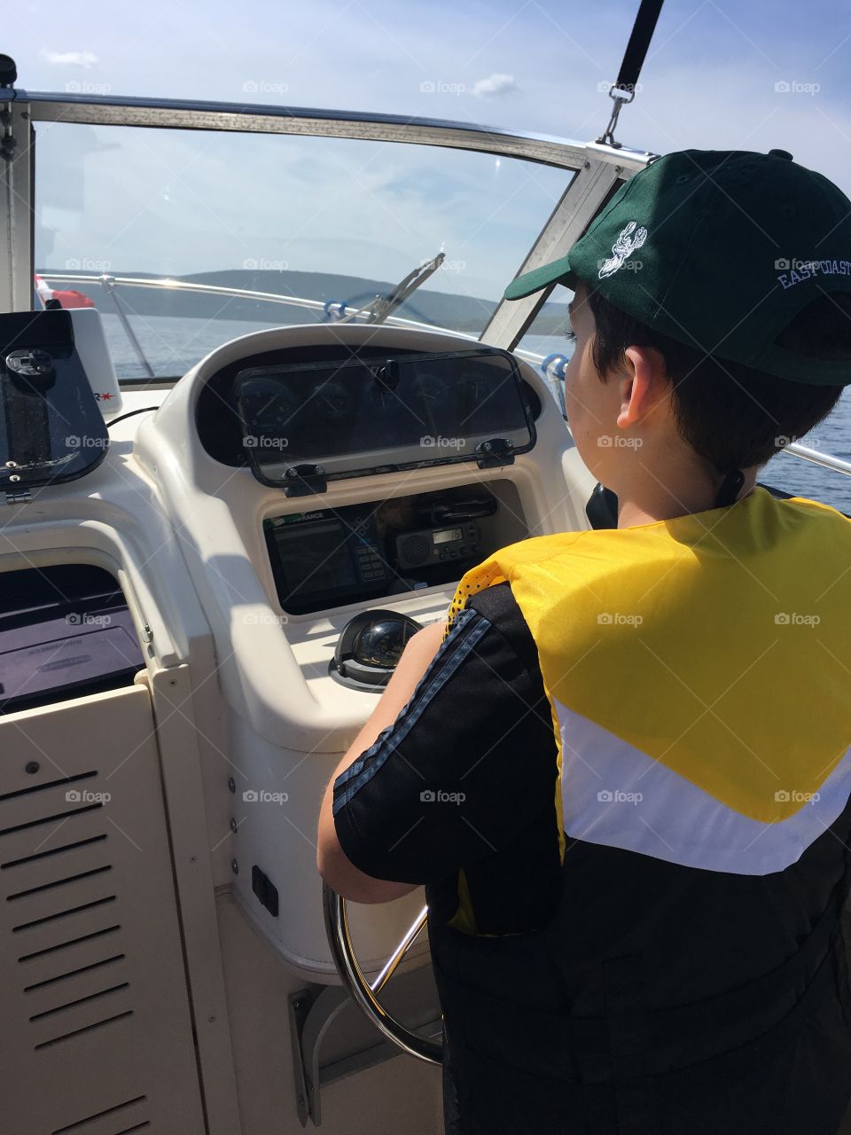 Young boy learning to drive a boat wearing a yellow life jacket and green ball cap. 