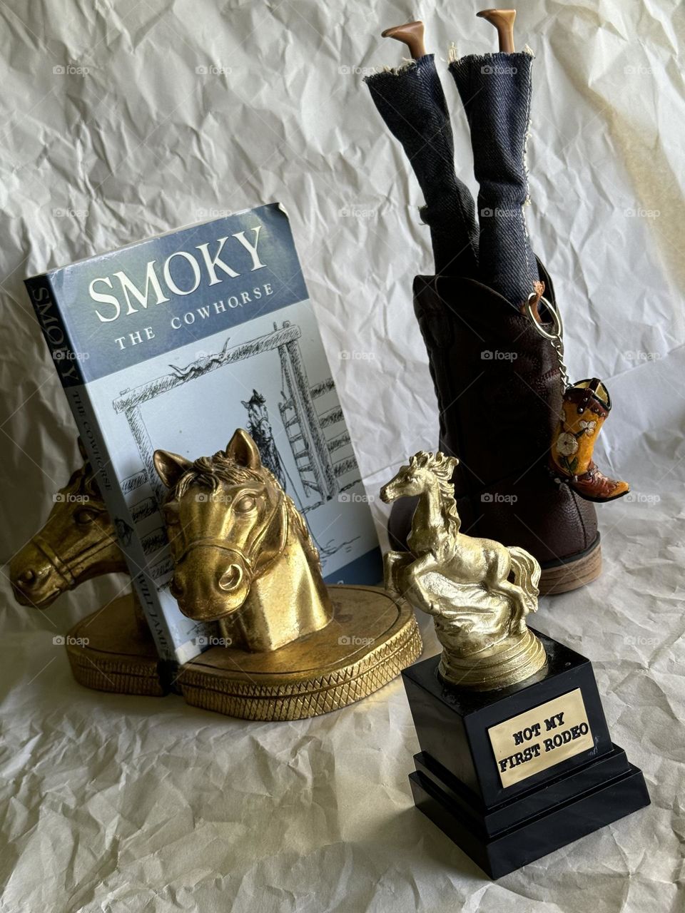 Western craze addressed with “Smoky the Cowhorse” (1926) between golden horse head bookends accompanied by blue jeaned doll stuffed in cowboy boot while holding decorated boot keychain with “Not My First Rodeo” trophy with rearing golden stallion