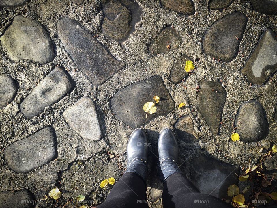 Looking down at woman’s black boots on pavement with fallen leaves beside