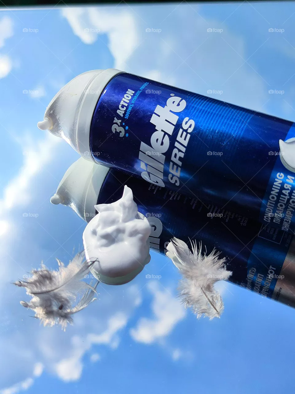 Men's cosmetics. On a mirrored surface lies a blue bottle with a white inscription: "Gillette series", two bird feathers and white fluffy foam. The mirror surface reflects objects on the mirror surface and the blue sky with white clouds