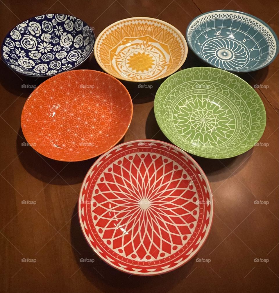 A vibrant set of ceramic bowls in complimentary colors—navy blue, light blue, red, orange, yellow, and green—arranged side by side, creating a cheerful, eye-catching rainbow of glossy finishes and bold tones.