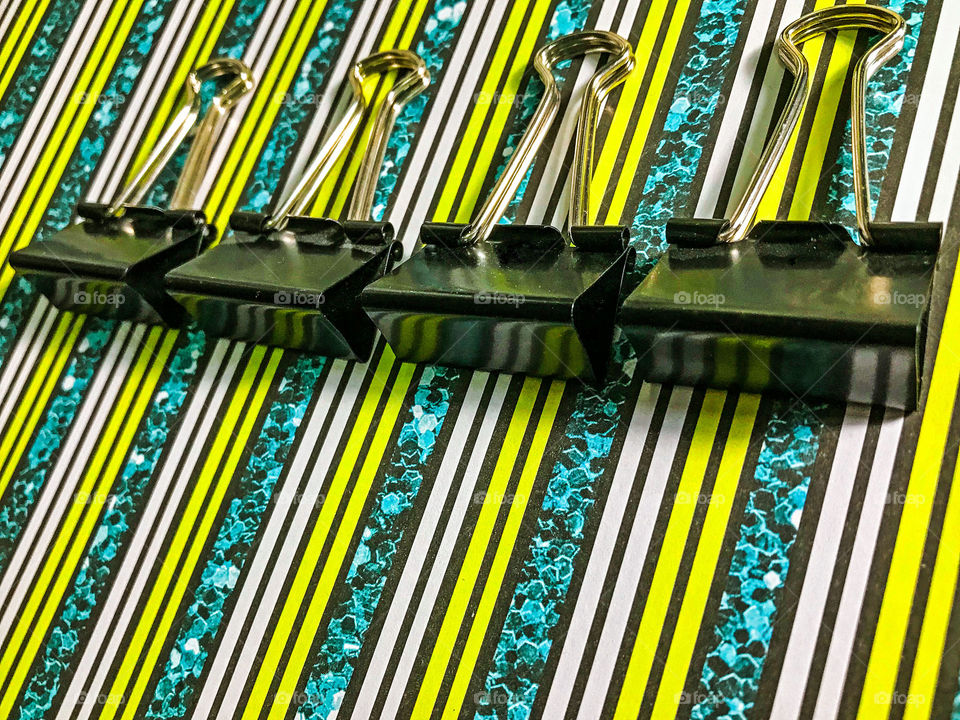 Black binder clips on striped paper. The flat rectangular part of the clip is reflecting the lines of the paper making more little rectangles in the reflection.